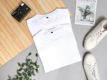 Flatlay photo featuring 2 t-shirts folded on the ground next to sneakers, plant,magazine and camera