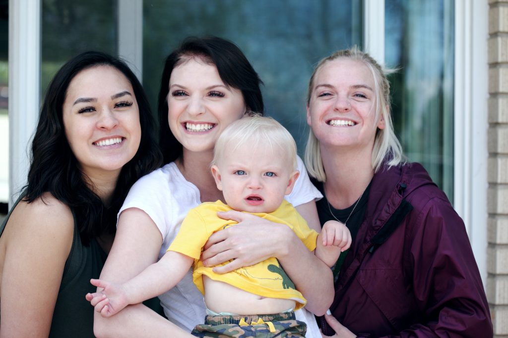 Three smiling young women in their twenties with a one year old child.