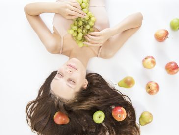 woman lying on white flooring holding grapes beside apple fruits